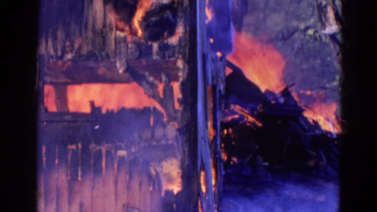 Flames burn through a wood-sided structure with smoke and charred openings.