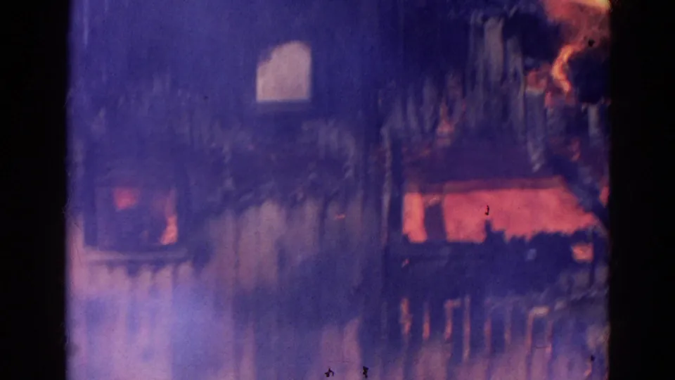 Soft, smoky 8mm view of the house fire with flames visible through openings in the damaged wall.