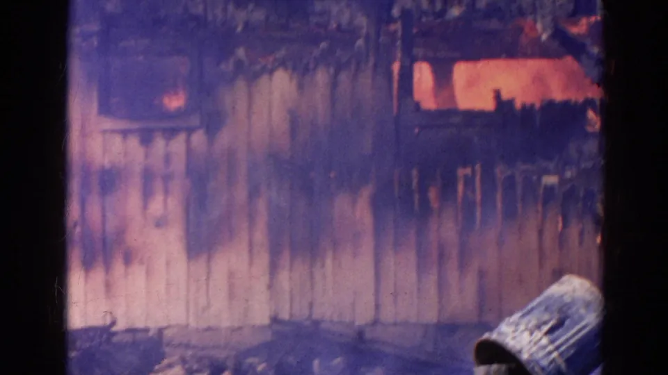 Late-clip frame from the Wauconda fire showing smoke, glowing flames, wood siding, and debris near the structure.