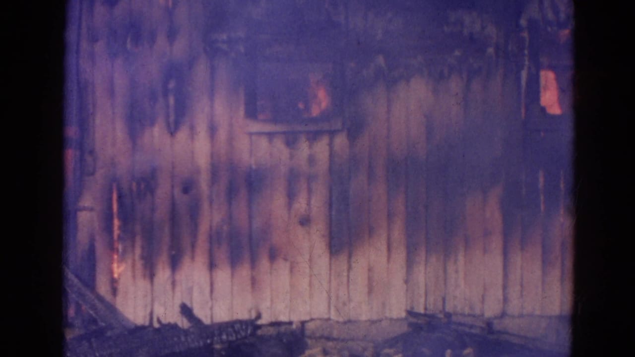 Close view of scorched wooden siding with dark smoke and faint orange firelight behind openings.