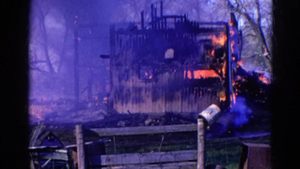 1968 Wauconda 8mm frame showing a burning wooden building beyond a fence, with smoke, trees, debris, and a small metal canister visible.