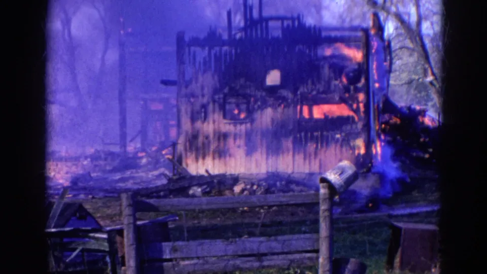 1968 Wauconda 8mm frame showing the structure still burning amid blue smoke, with charred siding, fence posts, trees, and debris.