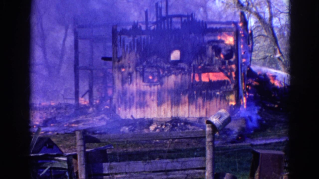 1968 Wauconda 8mm frame showing a wooden structure burning behind a fence, with smoke, flames, trees, and a mailbox visible.