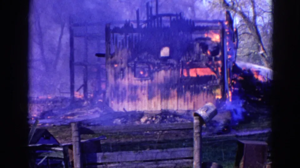 1968 Wauconda 8mm frame showing a wooden structure burning behind a fence, with smoke, flames, trees, and a mailbox visible.