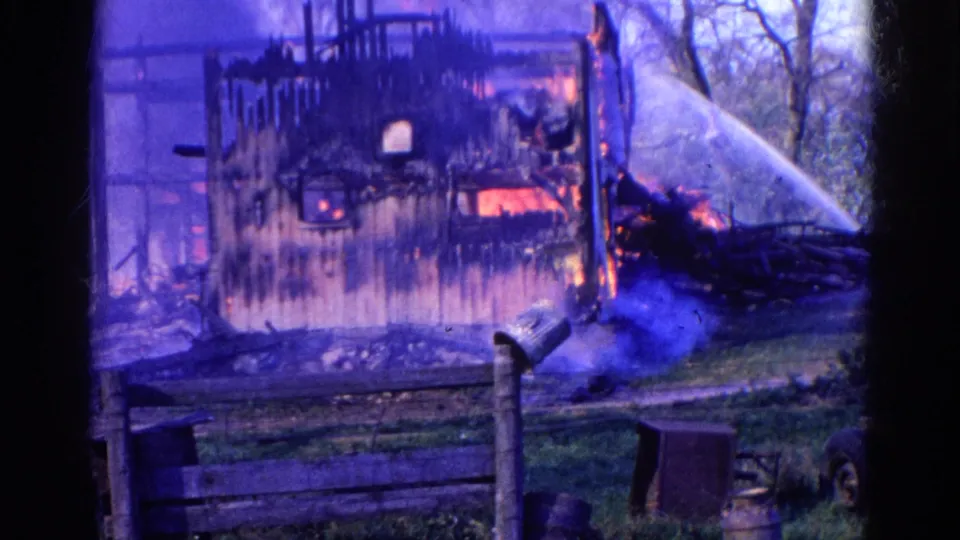 1968 Wauconda 8mm frame with water spraying across a yard toward a burning wooden structure behind a fence.