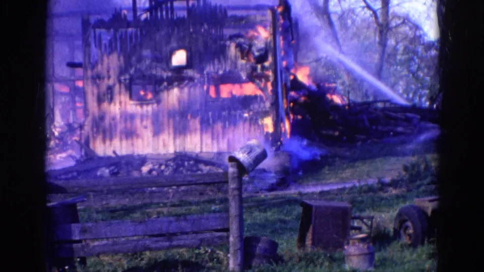 Final 1968 Wauconda frame showing the burning structure, hose spray, smoke, fence, mailbox, and nearby yard objects.
