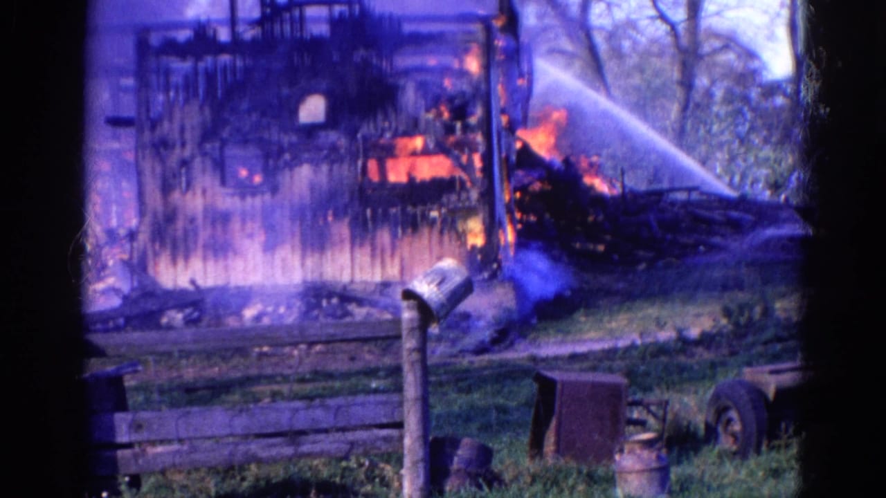 Burning barn with flames, smoke, hose spray, fencing, and yard objects in the foreground.