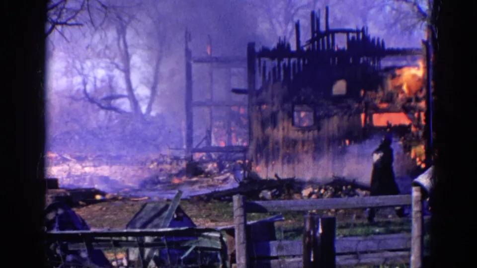 The 8mm frame shows smoke hanging over the burned barn structure as a firefighter stands near debris and a low fence.
