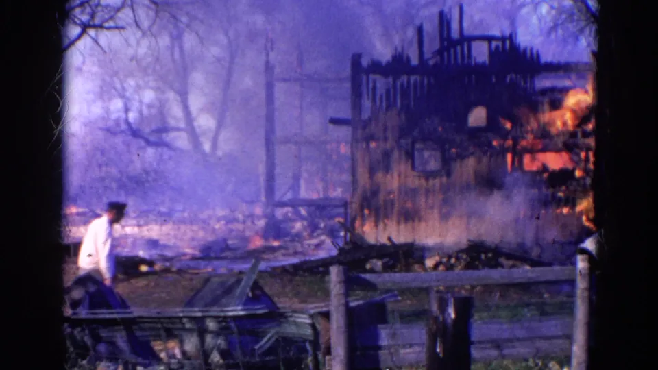 A bystander or responder in a white shirt moves near the foreground while the Wauconda barn continues burning behind smoke and debris.