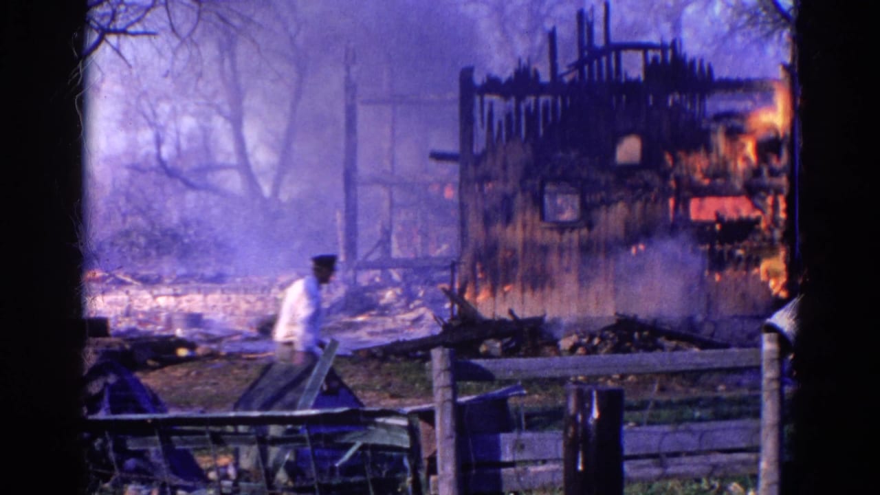 In 1968 Wauconda, Illinois 8mm footage, a man in a white shirt and dark hat passes a smoking, burning house behind a wooden fence.