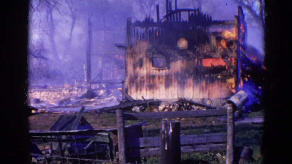 The 8mm frame shows the burned house shell in Wauconda with smoke in the trees and a firefighter hose spray beginning at the right edge.