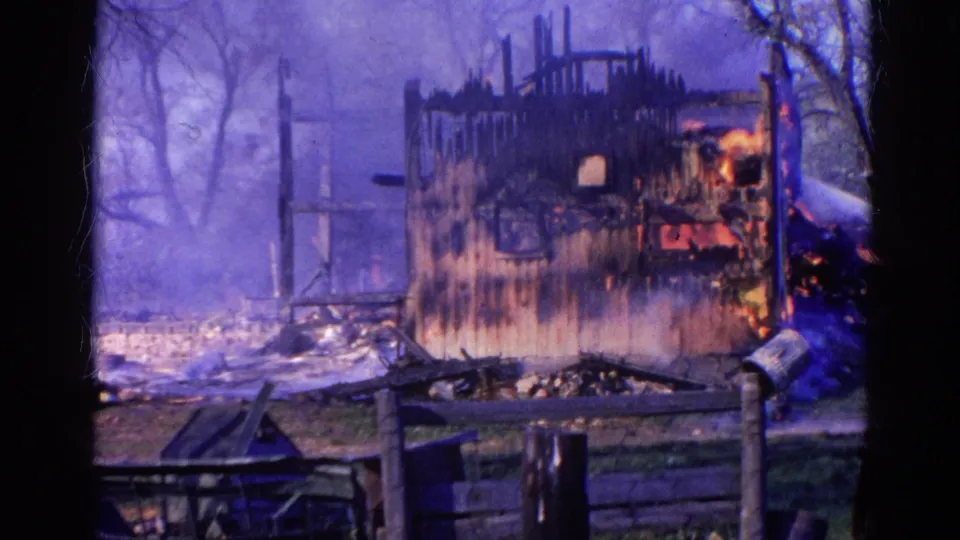 A hose stream sprays across the right side of the 1968 Wauconda house fire as smoke surrounds the charred wooden structure.