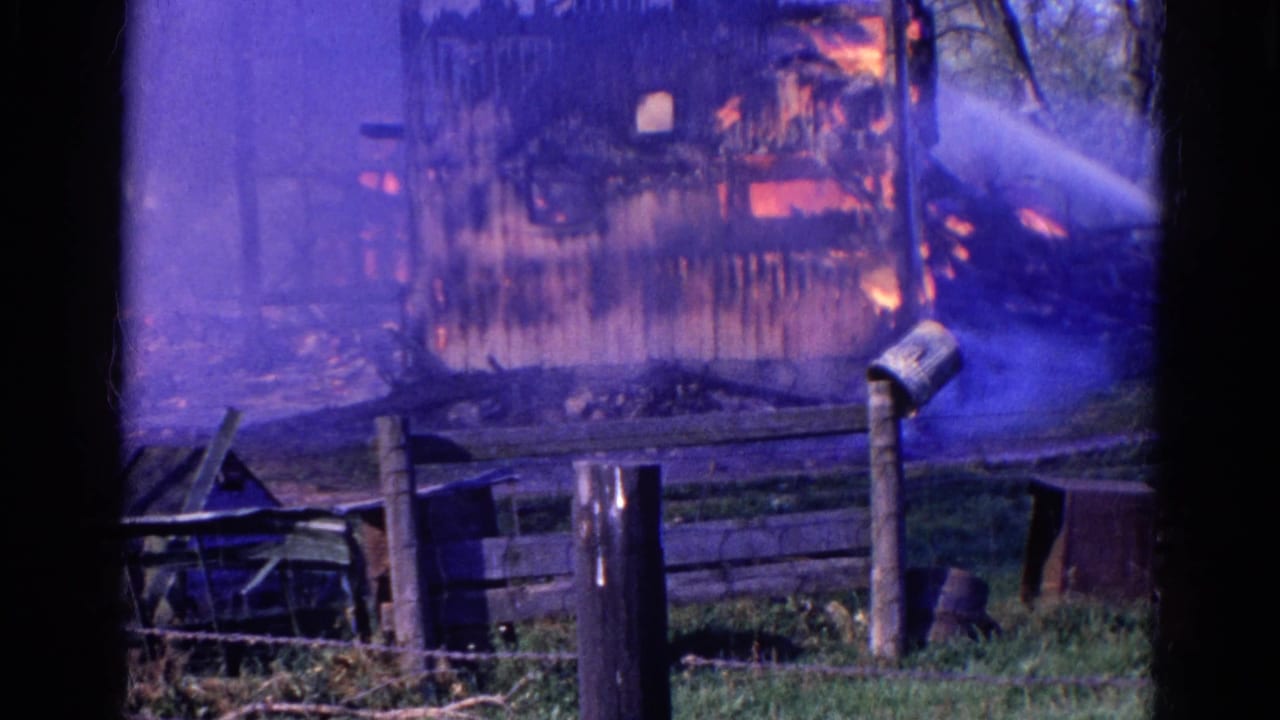 Water sprays onto a burning rural structure behind a fence and grassy foreground.