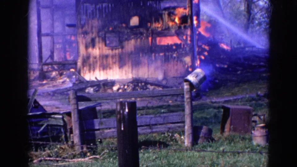 Late-1960s home-movie view of a Wauconda, Illinois structure fire framed by fence posts and grass.