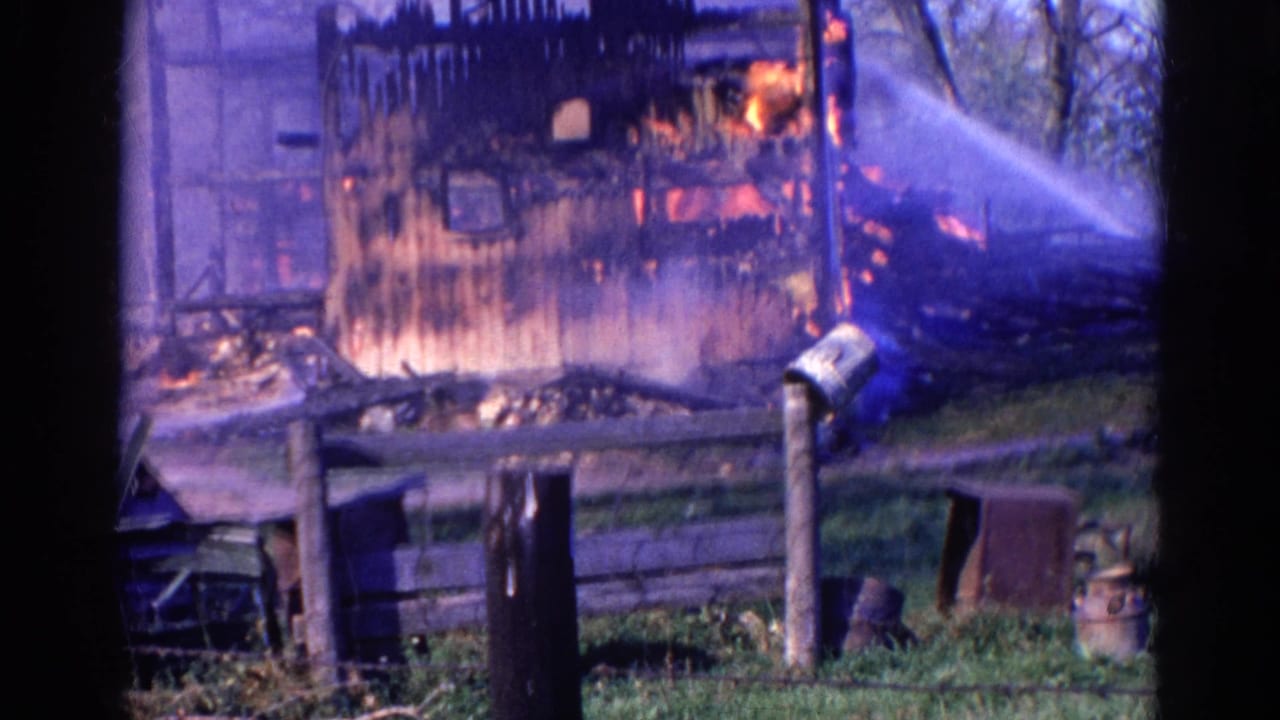 Burning wooden barn with water spray, smoke, fence, mailbox, and dark framing in the foreground.