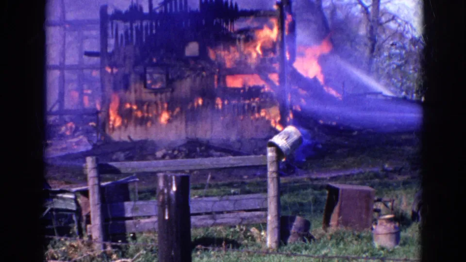 1968 Wauconda, Illinois frame with bright flames inside a partially burned barn and a firefighter hose stream cutting across the scene.