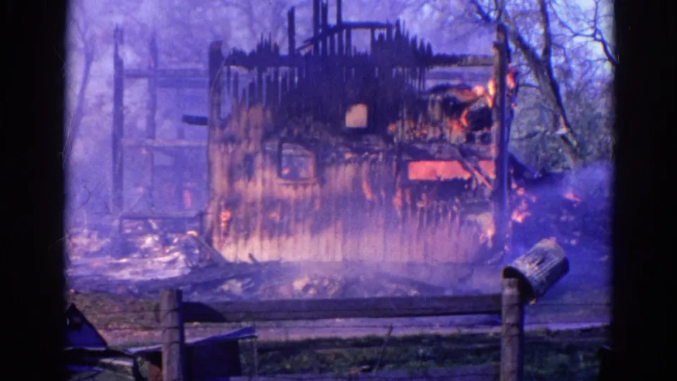 1968 Wauconda, Illinois 8mm still showing smoke surrounding a fire-damaged barn, with visible flames, fence rails, grass, and a roadside mailbox.