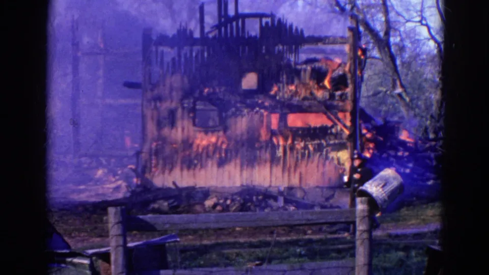 1968 Wauconda, Illinois home-movie frame of a charred barn still burning behind a fence, with smoke, flames, a mailbox, and dark foreground framing.