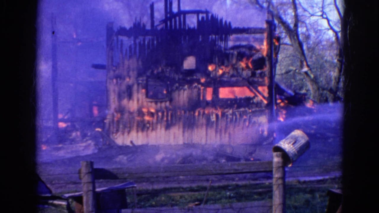 1968 Wauconda, Illinois 8mm frame showing a charred wooden house with smoke, flames, water spray, fence rails, and a roadside mailbox.