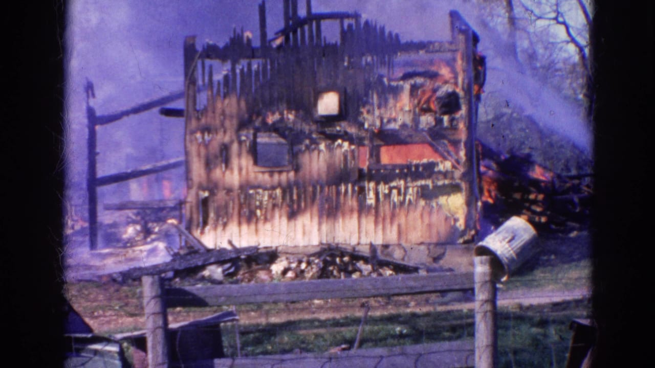 Charred house frame with smoke, burned debris, fence, mailbox, and water spray.