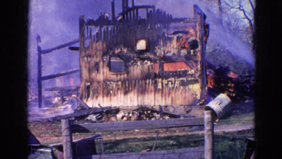 1968 Wauconda, Illinois frame of a heavily burned house shell with smoke, debris, fence rails, mailbox, and firefighting water spray.