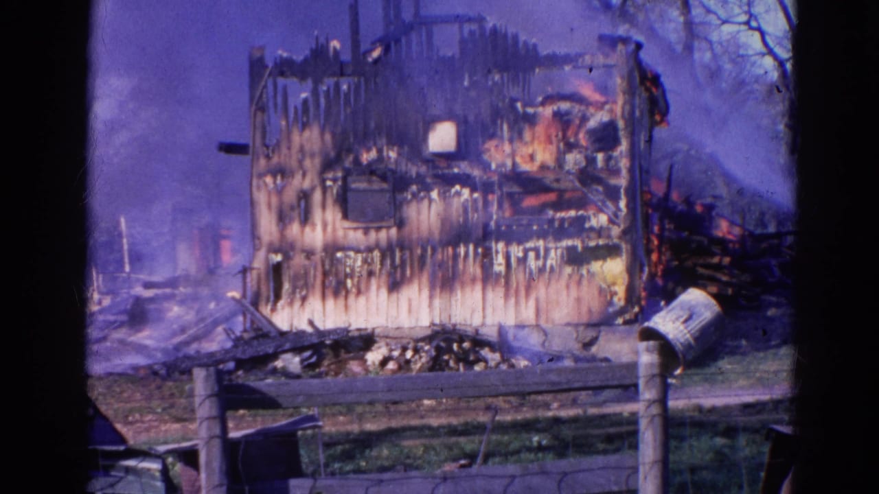 1968 Wauconda, Illinois frame showing the blackened side of a burned house, smoke, remaining fire glow, fence rails, and mailbox.