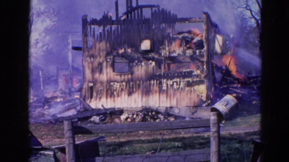 1968 Wauconda, Illinois frame of a fire-damaged house with smoke, visible flame on the right, foreground fence, mailbox, and scattered debris.