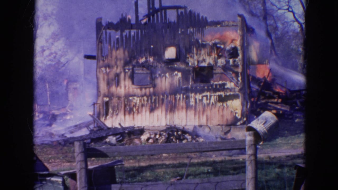 1968 Wauconda 8mm frame showing a charred house exterior with smoke, water spray, fence, grass, and leafless trees.