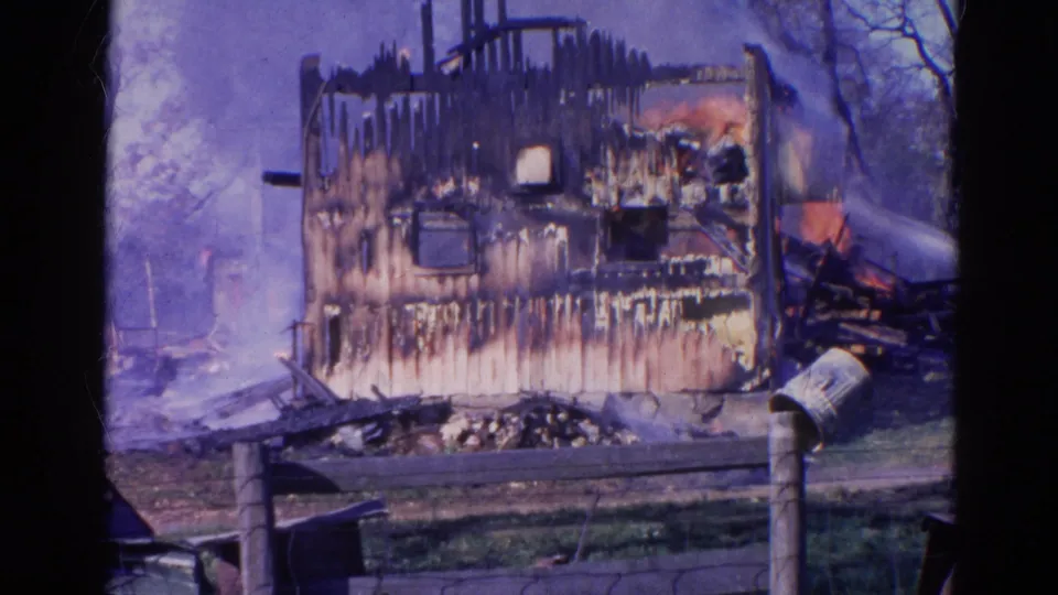 1968 Wauconda 8mm frame showing a charred house exterior with smoke, water spray, fence, grass, and leafless trees.