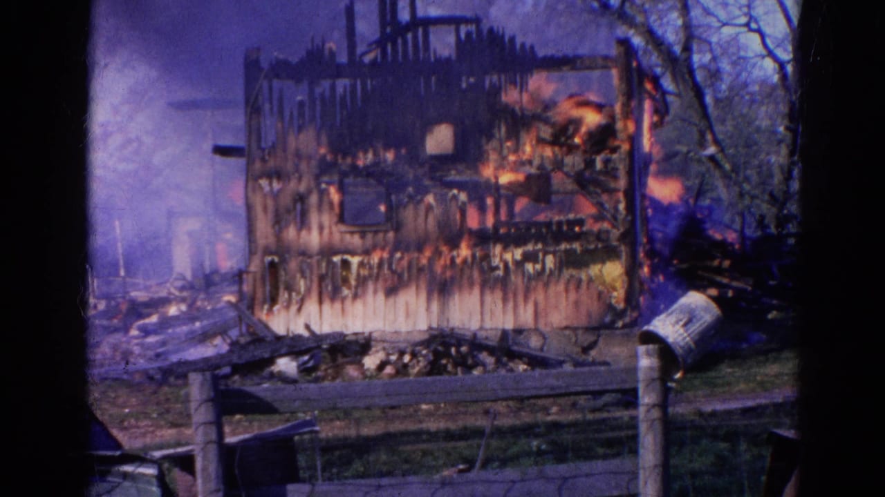 Flames burn inside a damaged house wall while smoke and water spray cross the scene.