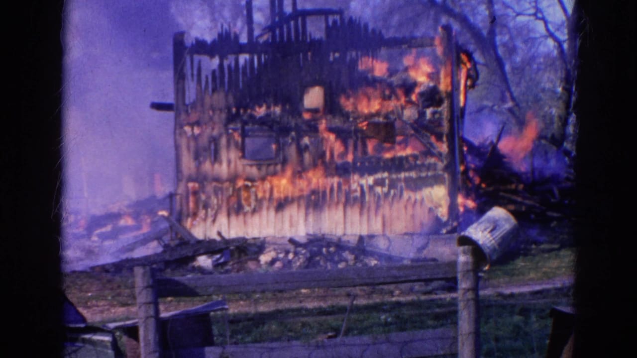 1968 Wauconda home-movie frame showing flames spreading across a charred house exterior behind a low fence.