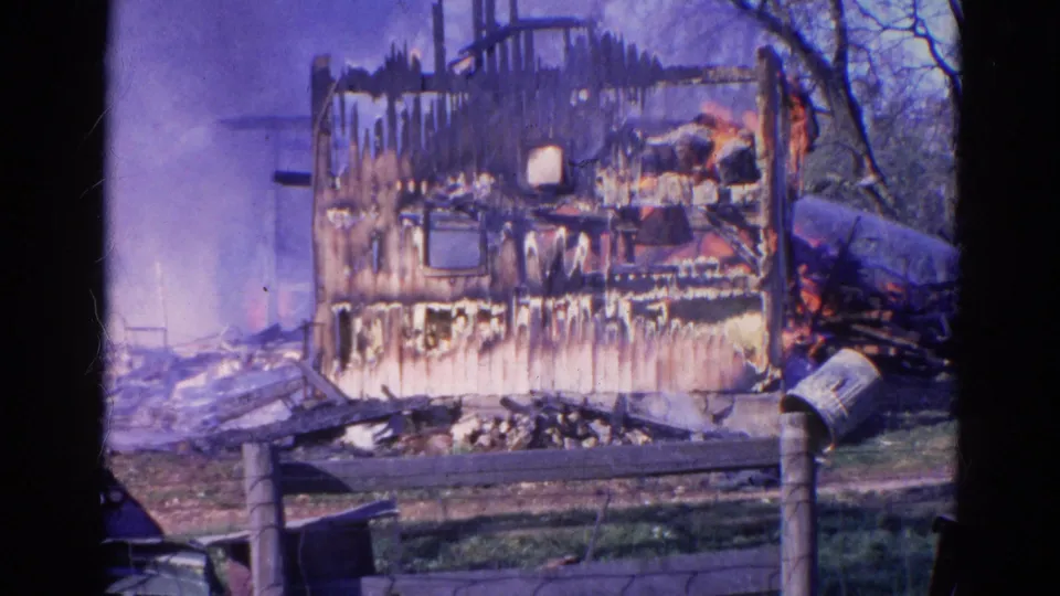 1968 Wauconda 8mm frame showing a heavily burned house wall with smoke, water spray, fence, grass, and bare trees.