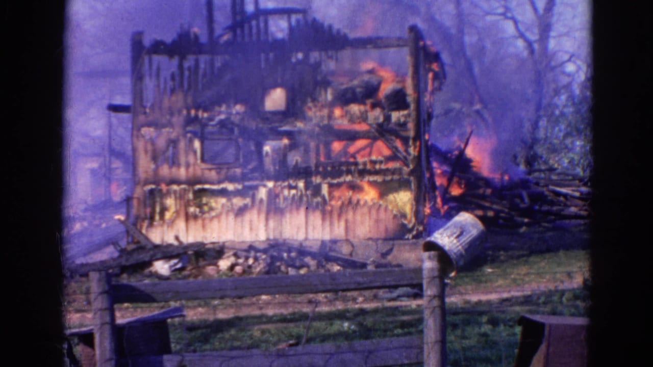 Wauconda, Illinois 1968 home-movie frame of flames inside a heavily damaged wooden structure with smoke and a fence in the foreground.