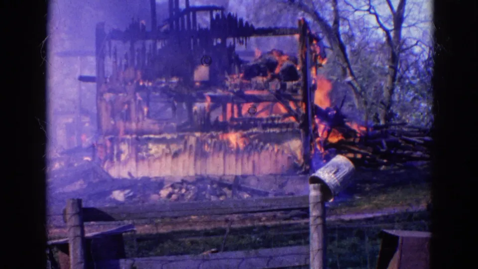 Vintage 8mm frame from Wauconda, Illinois showing a charred structure with active flames, smoke, fence posts, and trees nearby.