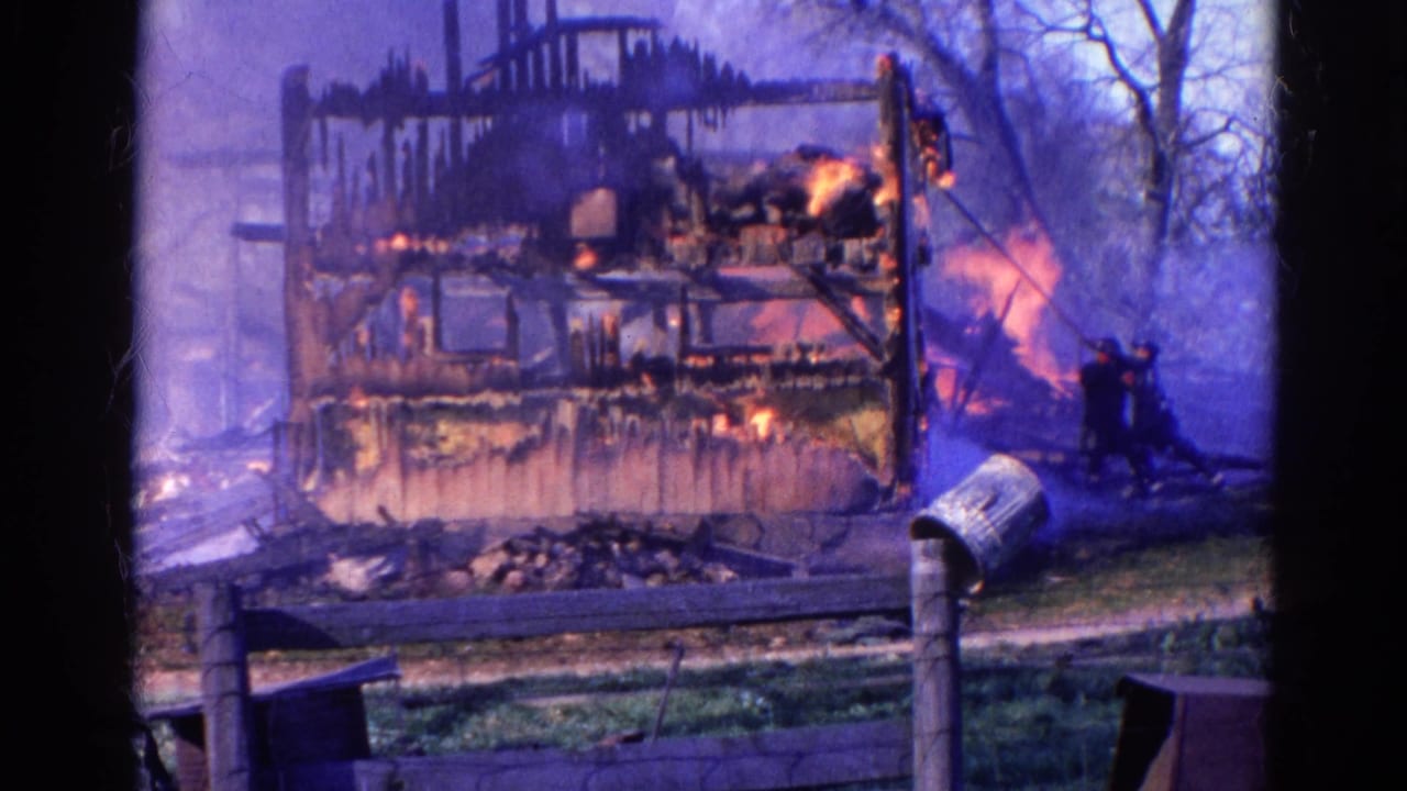 Charred house frame burns behind a fence with smoke, flames, two men, trees, and a metal can in front.
