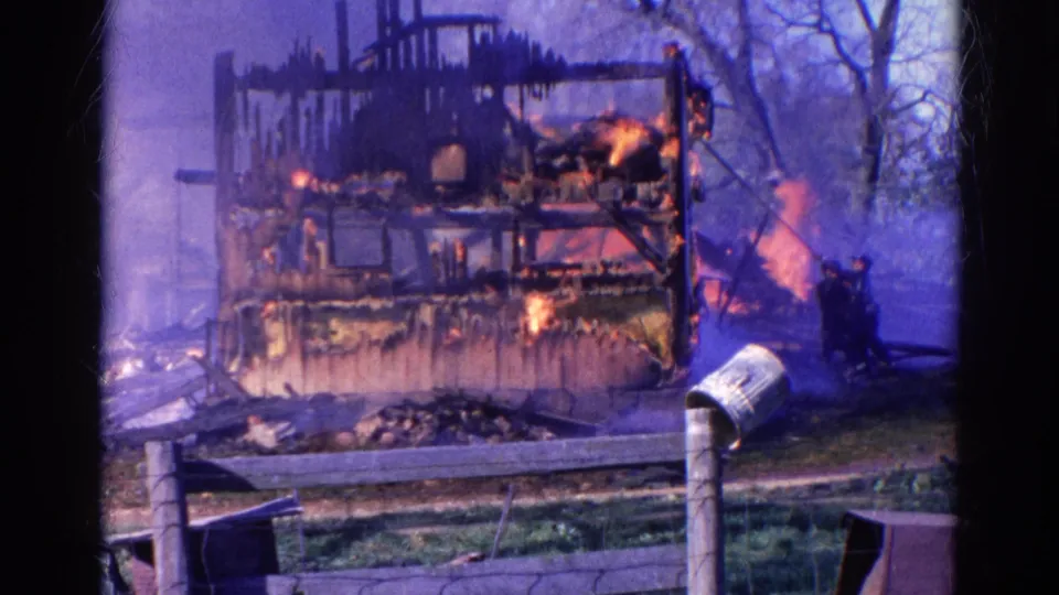 1968 Wauconda frame of a house fire viewed across a fence, with two men near the flames and smoke around the damaged frame.