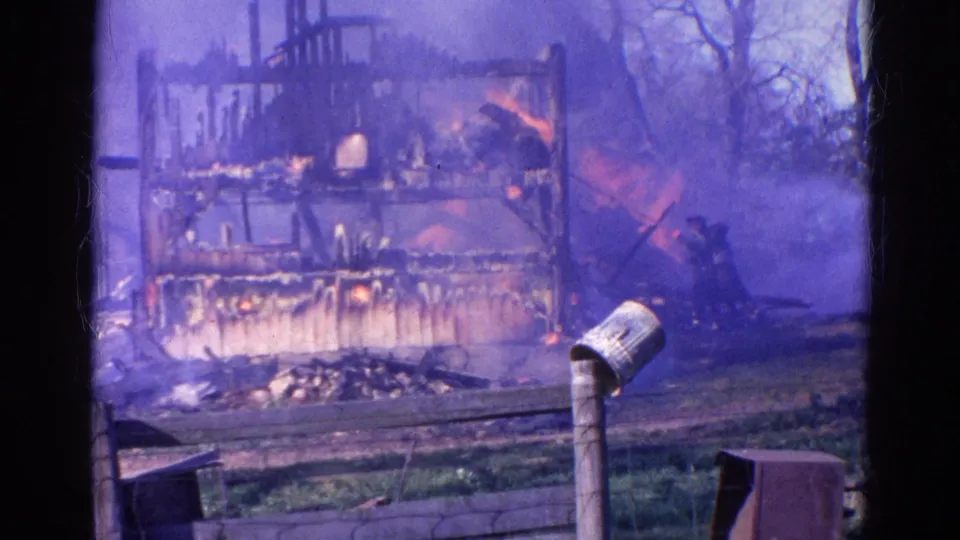1968 Wauconda frame showing the house fire through heavy smoke, with a man near the right side and fence details in front.
