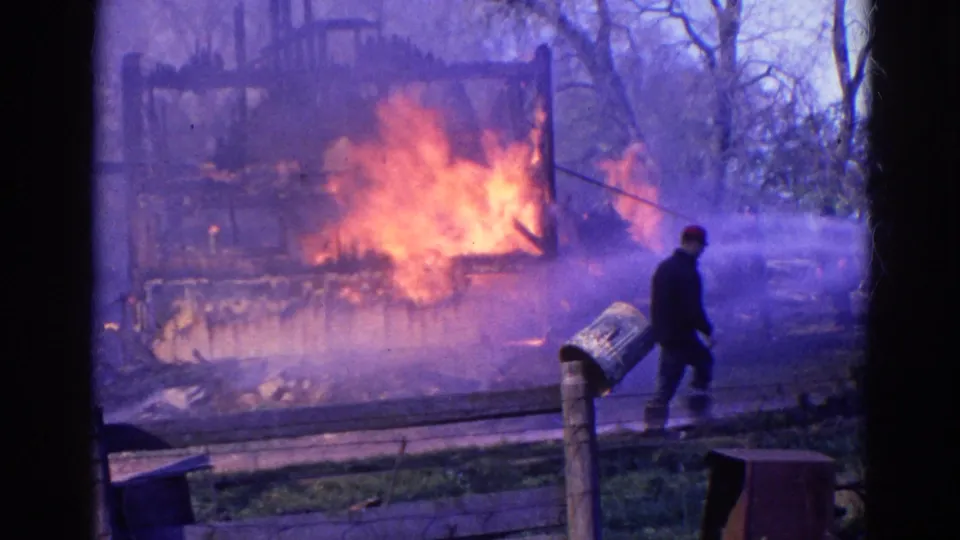 1968 Wauconda, Illinois 8mm frame showing an adult with a hose approaching a burning wooden structure amid smoke.