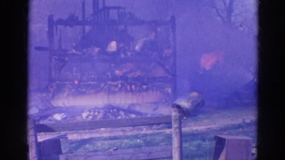 Smoke obscures the 1968 Wauconda fire scene, with the burning structure, fence, grass, and metal post still partly visible.