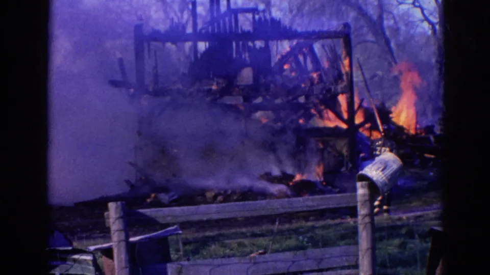 1968 Wauconda, Illinois 8mm frame showing flames along a damaged wooden structure, with smoke drifting across a fenced yard.