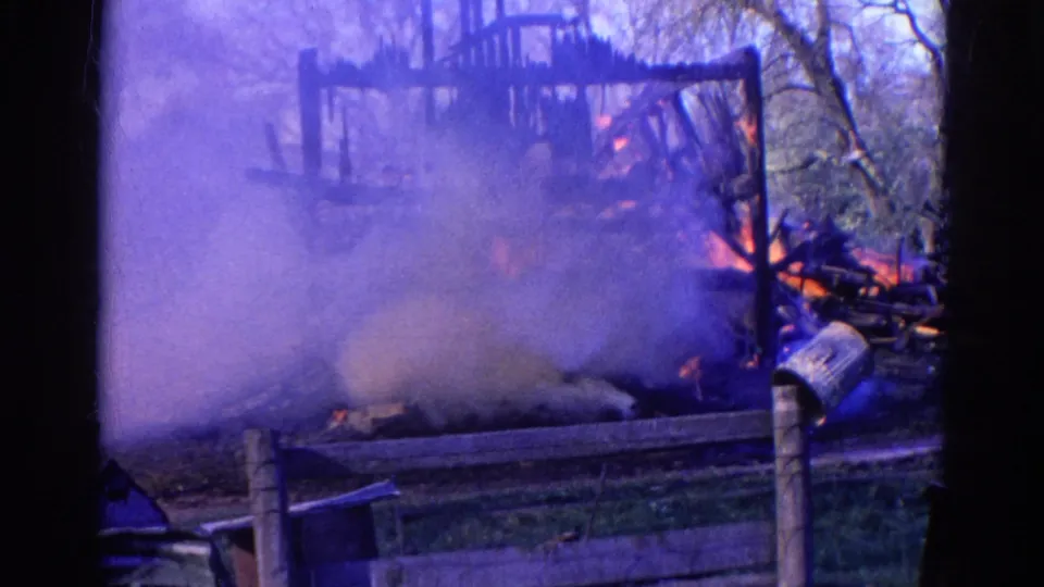 1968 Wauconda, Illinois 8mm frame showing smoke, flames, and a partially collapsed wooden structure behind a fence.