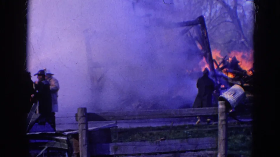 1968 Wauconda, Illinois fire footage frame showing figures, dense smoke, flames, and the remains of a wooden structure.