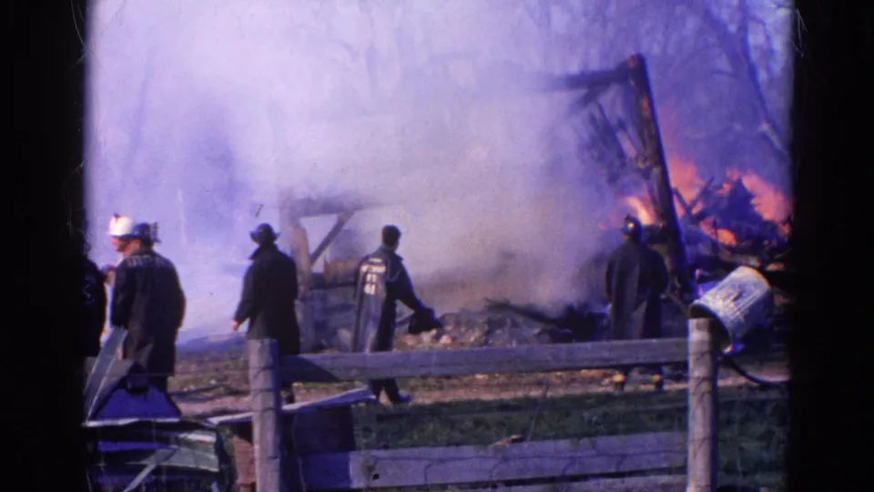 1968 Wauconda, Illinois frame with firefighters moving near a fence, smoke, burned debris, and flames at the right side.