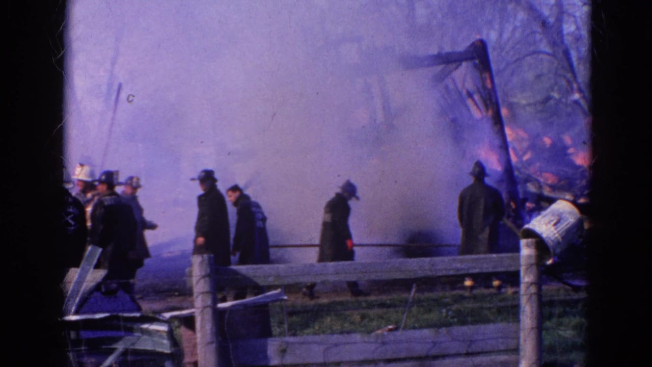 Firefighters and onlookers stand near heavy smoke behind a wooden fence.