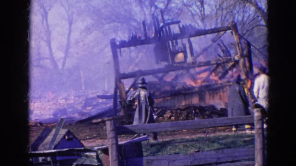 1968 home-movie frame of firefighters watching or working near a collapsed, burning barn structure behind a rural fence.