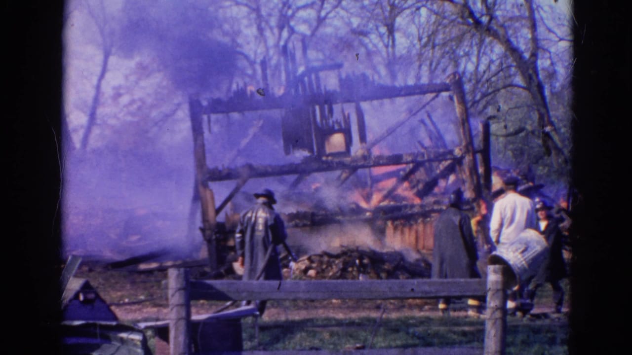 Flames and smoke rise from a wooden structure while people stand near a fence.