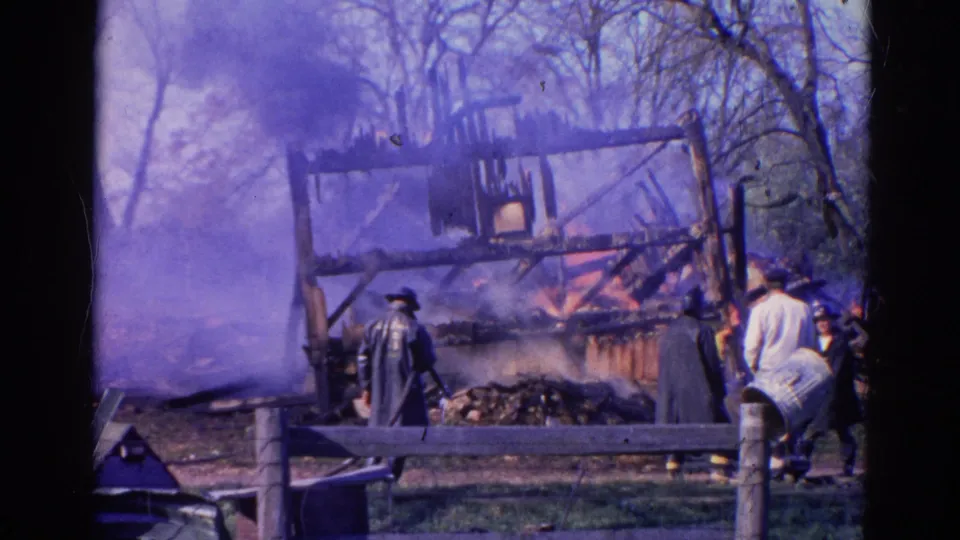 1968 Wauconda home-movie frame showing a burning wooden structure, smoke, bystanders, and possible firefighters near a fence.