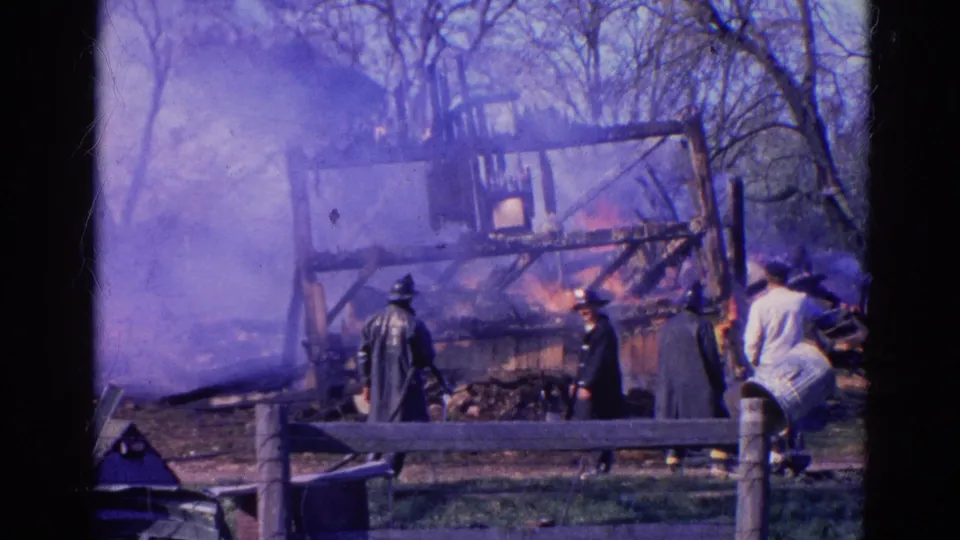 1968 Wauconda frame with flames, smoke, a charred wooden frame, bystanders, and a dark-uniformed figure near the fire.