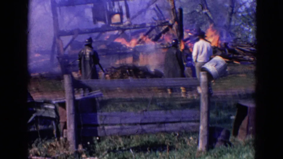 Home-movie view from Wauconda in 1968 showing smoke, fire, a collapsed wood frame, fence rails, and nearby observers.