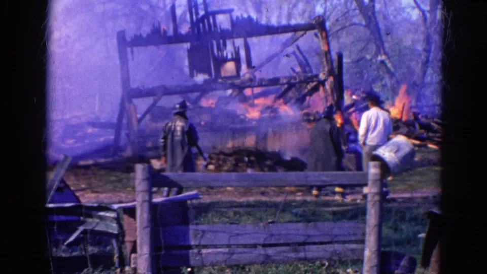 1968 Wauconda 8mm frame with a wider view of a burning wooden frame, smoke, bystanders, fence posts, and trees.